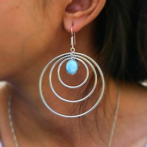 May include: Silver hoop earrings with a light blue oval gemstone at the center. The earrings feature three concentric hoops of decreasing size, creating a layered effect. The hook is a simple wire design.