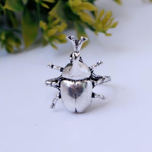 May include: A silver-toned ring featuring a detailed rhinoceros beetle design. The beetle has a prominent horn, textured legs, and a smooth, rounded body. The metal has a polished finish with darkened recessed areas to highlight the insect's anatomy.
