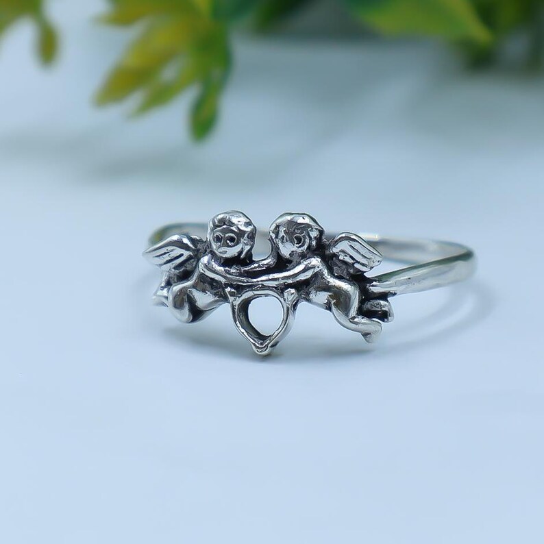 May include: A silver-toned ring featuring two cherub figures facing each other with wings extended. The cherubs are connected at the center, forming a small heart-shaped cutout between their lower bodies. The band is smooth and polished silver.