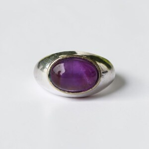 May include: A silver ring featuring a large, oval, deep purple gemstone. The ring has a smooth, rounded band and a bezel setting that holds the gemstone securely. The polished gemstone reflects light, creating a striking appearance.