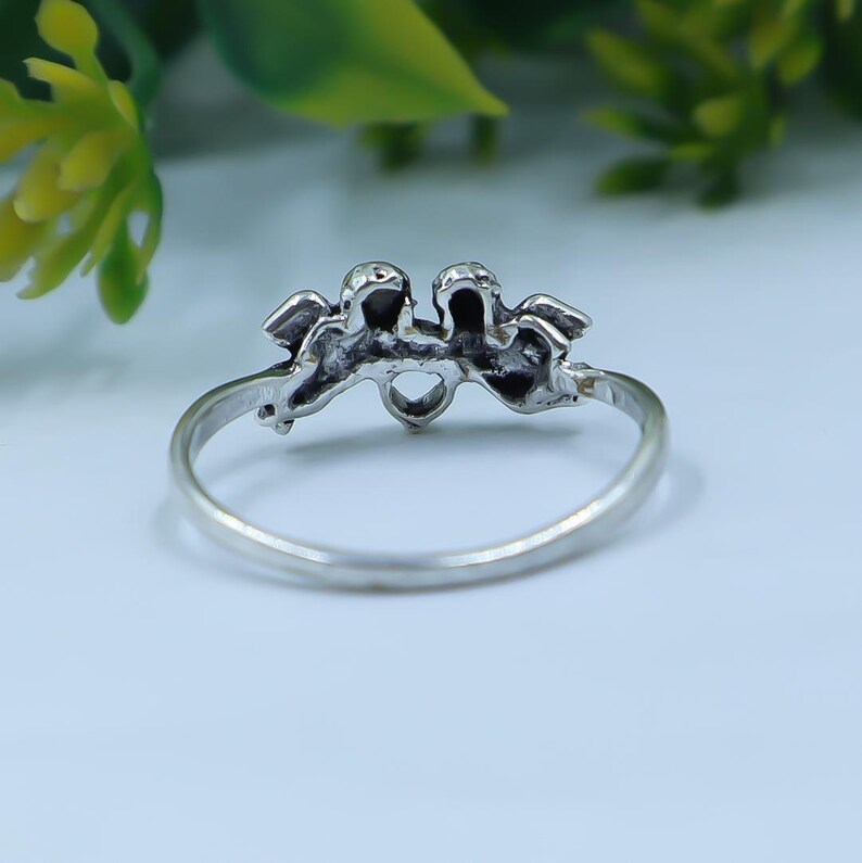 May include: A silver-toned ring featuring a decorative centerpiece shaped like two small figures or cherubs positioned side by side. The metal band is thin and circular, with the detailed ornament centered at the top of the ring's band structure.
