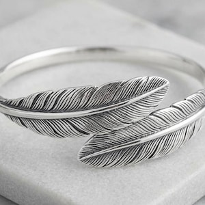 Pode incluir: Uma pulseira de punho prateada com dois desenhos detalhados de penas. As penas são intrincadamente esculpidas com linhas finas, criando uma aparência texturizada. A pulseira tem um acabamento polido e um design simples e elegante.