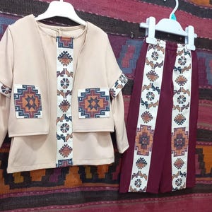 May include: A beige jacket and maroon trousers set featuring white vertical panels with geometric patterns in orange, blue, and brown. The jacket has two square pockets on the front, and both pieces are displayed on white plastic hangers.