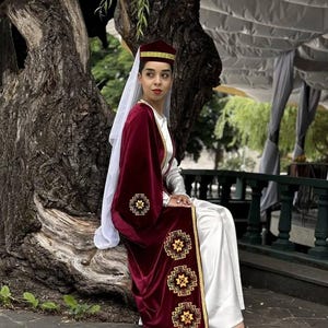 May include: A person wearing a traditional burgundy velvet robe with gold embroidery, a matching hat, and a white dress. The person is sitting near a large tree trunk, with a white veil and red shoes.