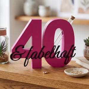 May include: A bright pink wooden number 40 decoration featuring a black cursive script overlay that reads & fabelhaft. The number zero includes a clear glass test tube insert with a cork stopper, designed to hold a single stem or small floral element.