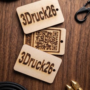 Könnte beinhalten: Drei rechteckige Holzanhänger mit dem Text "3Druck26" und einem QR-Code. Die Anhänger sind hellbraun mit dunkelbraun graviertem Text und Details. Die Anhänger liegen auf einer dunkelbraunen Holzoberfläche, mit schwarzen und goldenen Accessoires.
