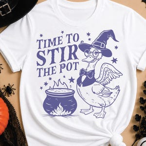 Peut inclure: Un t-shirt blanc avec un motif violet représentant un canard portant un chapeau de sorcière, des lunettes et un nœud papillon, à côté d'un chaudron bouillonnant. Le texte "TIME TO STIR THE POT" est entouré de petites étoiles violettes.