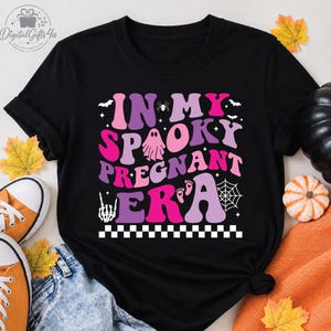 Peut inclure: Un t-shirt noir avec le texte "IN MY SPOOKY PREGNANT ERA" en lettres ondulées roses et violettes. Le motif comprend un petit fantôme, une araignée, une toile d'araignée, une main squelette et des empreintes de bébé, avec une bordure en damier noir et blanc.