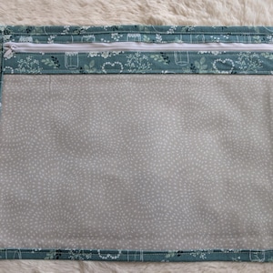 Pode incluir: Uma bolsa retangular com fecho de correr, apresentando uma borda de tecido azul-petróleo com uma estampa branca de plantas e frascos. O painel central é feito de material transparente com um padrão sutil de pontos brancos e fecho.