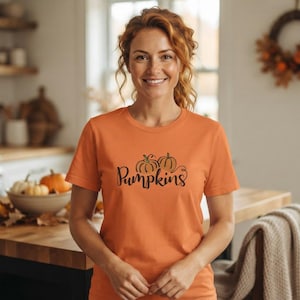 Pode incluir: Uma t-shirt de manga curta cor de laranja com um gráfico de duas abóboras em tons de laranja e verde, com a palavra Pumpkins escrita num tipo de letra cursivo preto centrado na zona do peito da peça de vestuário.