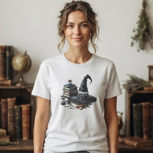 Pode incluir: Uma t-shirt de manga curta cinzento-claro com uma impressão gráfica de uma pilha de livros antigos encimada por um chapéu de bruxa preto pontiagudo e uma pequena garrafa de vidro. O design está centrado no peito da peça de vestuário.