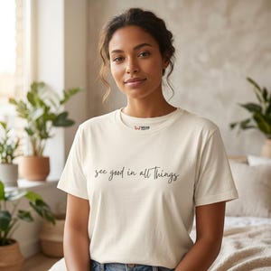 Pode incluir: T-shirt creme com a frase "see good in all things" em itálico. A camiseta de manga curta tem gola redonda e um logotipo da marca no peito. A camiseta é de cor neutra.