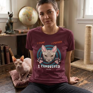 Pode incluir: Camiseta bordô com o texto "I CAME... I PURRED... I CONQUERED" e um gráfico de um gato Sphynx. O gato é sem pelos, com orelhas rosas e rosto cinza claro. Um gato Sphynx real está sentado em uma almofada.