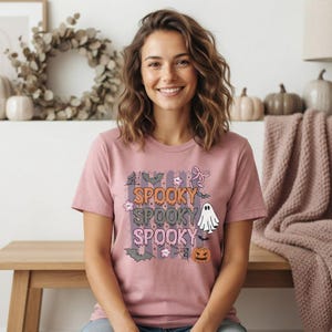 Pode incluir: Uma t-shirt de manga curta malva com uma impressão gráfica com o texto SPOOKY repetido três vezes em laranja e cinzento. O design inclui ilustrações de um fantasma branco, uma abóbora laranja, morcegos, teias de aranha e laços cor-de-rosa.