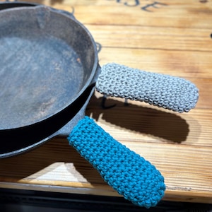 May include: A cast iron skillet with two crocheted handle covers. One handle cover is teal, and the other is gray. The skillet sits on a wooden surface.