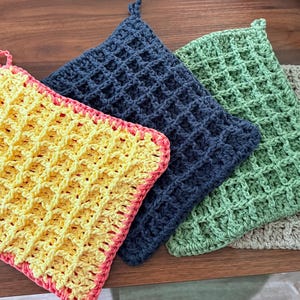 May include: Four square crochet dishcloths featuring a waffle stitch pattern. The set includes one yellow cloth with a pink border, one dark blue cloth, one light green cloth, and one beige cloth. Each dishcloth has a small loop for hanging.