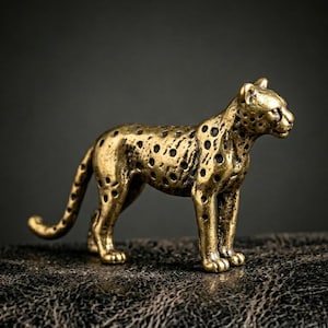 May include: A detailed brass leopard figurine with black spots. The sculpture is posed in a standing position, with its tail curved. The leopard is a golden color and is set against a dark background.