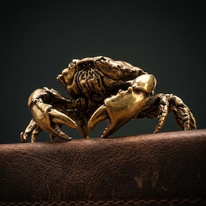 Könnte beinhalten: Detaillierte Krabbenfigur aus Messing mit strukturierten Oberflächen und markanten Scheren. Die Krabbe befindet sich auf einer braunen Lederoberfläche vor einem dunklen Hintergrund. Das goldene Aussehen deutet auf ein dekoratives Objekt hin.