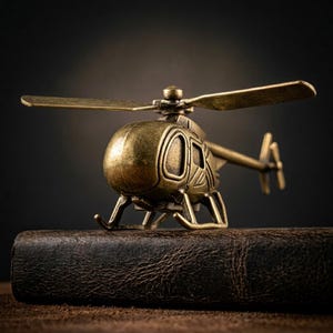 Puede incluir: Un detallado modelo de helicóptero de latón de estilo antiguo con aspas giratorias. El avión en miniatura se asienta sobre una superficie de cuero marrón oscuro texturizado, sobre un fondo oscuro borroso. El diseño del helicóptero incluye una cabina y un rotor de cola.