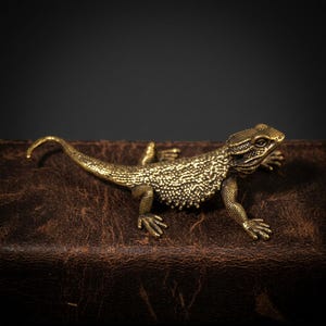 May include: A detailed brass lizard figurine with textured skin and a curled tail. The reptile is posed on a brown leather surface, with a dark background. The lizard's legs are outstretched, and its head is turned slightly.
