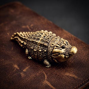 May include: A detailed, antique-style brass alligator figurine. The sculpture features textured scales and a detailed head with small, dark eyes. The alligator is resting on a brown leather surface, with a dark background.