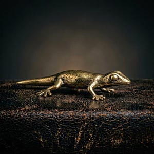 Puede incluir: Figura detallada de lagarto de latón con superficie texturizada. El lagarto está en posición de arrastrarse, con las patas extendidas y la cola arrastrando. La figura se encuentra sobre un fondo oscuro y texturizado.