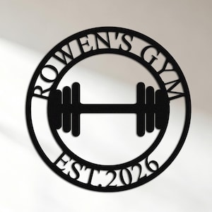 May include: A circular black wall sign featuring a barbell silhouette in the center. The text "ROWEN'S GYM" is arched along the top inner edge of the circle, and "EST. 2026" is arched along the bottom inner edge, all cut from a single piece of material.