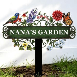 Puede incluir: Un letrero de jardín rectangular verde sobre una estaca negra con el texto blanco NANA'S GARDEN. El letrero está decorado con un pájaro pintado de colores, una mariposa monarca y varios elementos florales que incluyen flores rojas, moradas y blancas.