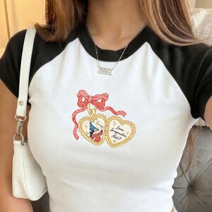 May include: White crop top with black raglan sleeves featuring a heart-shaped graphic with a cartoon character and the text "Some Imagination Huh?". The top is paired with gray sweatpants and a white purse.