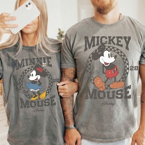 Puede incluir: Camisetas grises con Mickey y Minnie Mouse. Las camisetas tienen un dise&ntilde;o vintage con la imagen del personaje dentro de un marco circular. Los textos "Mickey Mouse" y "Minnie Mouse" est&aacute;n impresos debajo.