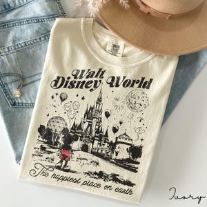 May include: Ivory-colored t-shirt with a black graphic of Mickey Mouse in front of a castle, with the words "Walt Disney World" and "The happiest place on earth." The shirt is paired with light blue jeans, a tan hat, and a necklace.