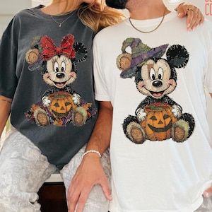 May include: A charcoal gray t-shirt featuring a graphic of a stitched-style mouse with a red bow and a pumpkin, and a white t-shirt featuring a graphic of a stitched-style mouse with a purple hat and a pumpkin, both labeled COMFORT COLORS.