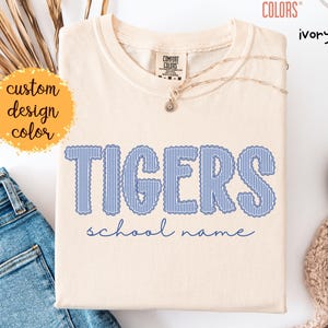 May include: An ivory t-shirt featuring the word TIGERS in blue and white vertical stripes with a scalloped edge, and the text school name in blue cursive script below. The shirt has a Comfort Colors brand label at the inner neckline.