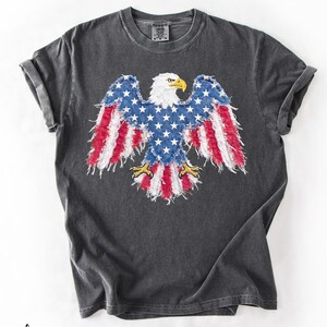 May include: Dark gray t-shirt featuring a distressed graphic of an American bald eagle with wings designed as the American flag. The eagle has a white head, yellow beak and talons, and blue wings with white stars and red stripes.