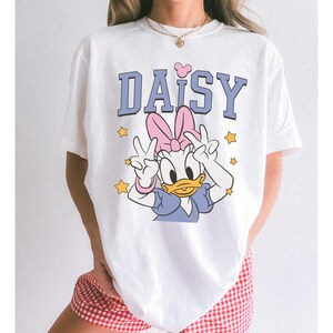 以下が含まれることがあります： デイジーダックのグラフィックがプリントされた白いTシャツ。「DAISY」の文字は青色のブロック体で、ピンクのリボンとピースサインをするデイジーダックの漫画のイメージが描かれています。黄色の星がグラフィックを囲んでいます。