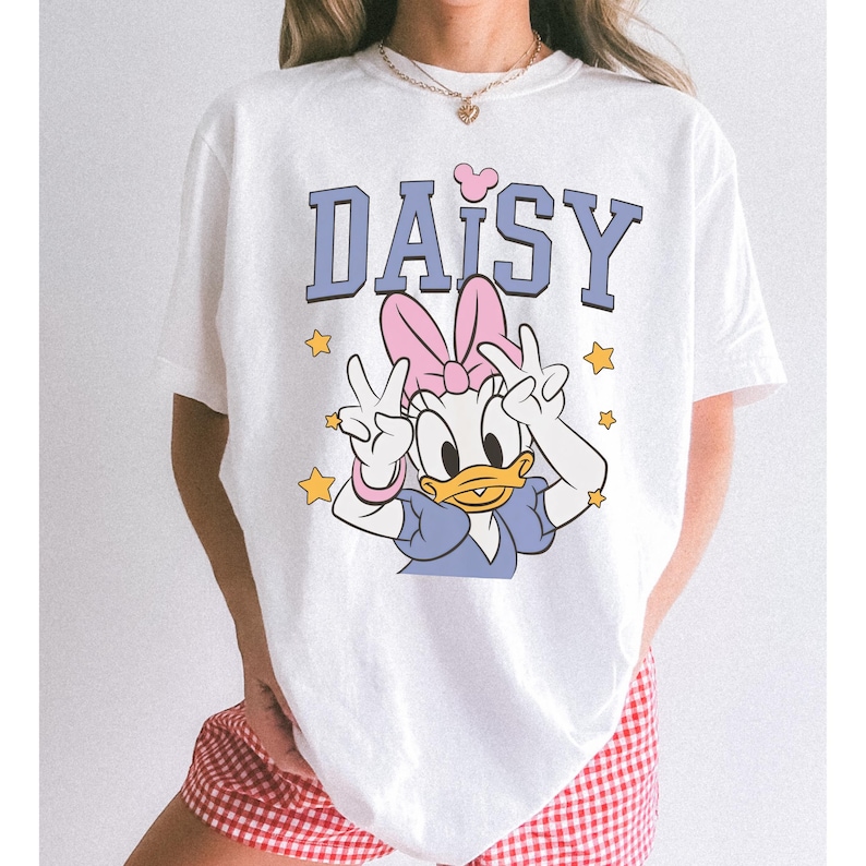 以下が含まれることがあります： デイジーダックのグラフィックがプリントされた白いTシャツ。「DAISY」の文字は青色のブロック体で、ピンクのリボンとピースサインをするデイジーダックの漫画のイメージが描かれています。黄色の星がグラフィックを囲んでいます。