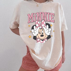 以下が含まれることがあります： ミニーマウスのグラフィックが特徴のクリーム色のTシャツ。「ミニー」の文字がキャラクターの上にピンク色でプリントされています。ミニーは両手でピースサインをしています。黄色の星がキャラクターを囲んでいます。