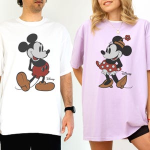 Puede incluir: Camisetas blancas y lavanda con Mickey y Minnie Mouse. Mickey lleva pantalones cortos rojos y zapatos marrones, mientras que Minnie lleva una falda de lunares rojos y zapatos marrones. Ambas camisetas tienen el logo de Disney.