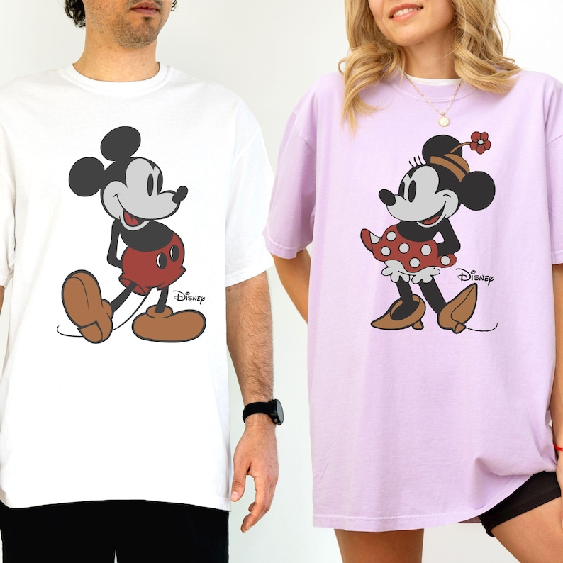 Puede incluir: Camisetas blancas y lavanda con Mickey y Minnie Mouse. Mickey lleva pantalones cortos rojos y zapatos marrones, mientras que Minnie lleva una falda de lunares rojos y zapatos marrones. Ambas camisetas tienen el logo de Disney.