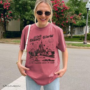 Puede incluir: Camiseta color carmes&iacute; con un gr&aacute;fico negro del castillo de Walt Disney World, fuegos artificiales y globos aerost&aacute;ticos. El texto "Walt Disney World" y "The happiest place on earth" est&aacute;n impresos en la camiseta. La camiseta es de manga corta y cuello redondo.