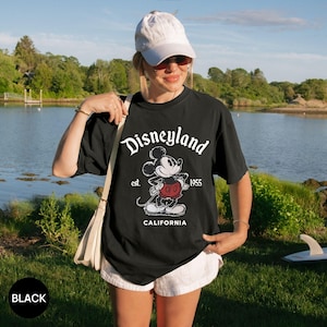 May include: Black t-shirt with a Disneyland graphic featuring Mickey Mouse. The design includes the text "Disneyland" in a script font, "est. 1955", and "CALIFORNIA". The shirt is a classic, vintage-style design.
