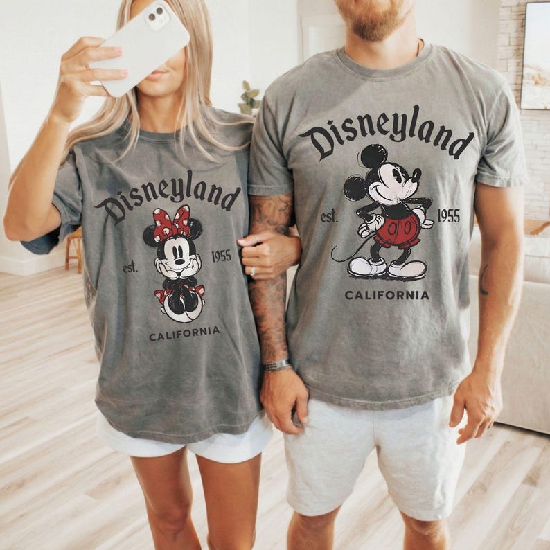 May include: Gray t-shirts with Disneyland graphics. One shirt features Minnie Mouse with a red bow, the other Mickey Mouse. Both shirts have the text "Disneyland est. 1955 CALIFORNIA".