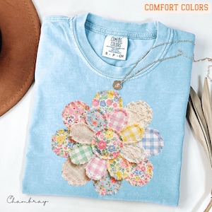 以下が含まれることがあります： 花柄アップリケのデザインが施された水色のTシャツ。アップリケは、ピンク、黄色、青、緑の様々な柄の生地で作られています。Tシャツには「Comfort Colors」と「Chambray」の文字が入っています。シルバーのネックレスも写っています。