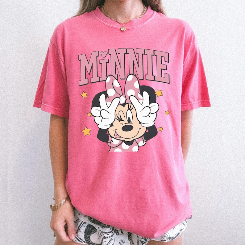 以下が含まれることがあります： ミニーマウスのグラフィックが特徴のピンク色のTシャツ。「MINNIE」の文字がピンク色のブロック体で描かれ、ミニーマウスがピースサインをしています。黄色の星がキャラクターを囲んでいます。