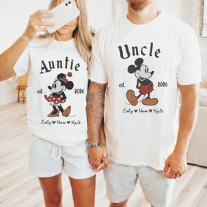 May include: Matching white t-shirts with Mickey and Minnie Mouse designs. One shirt says "Auntie" and the other says "Uncle", both with the year "2026" and names below. The shirts are paired with gray shorts.
