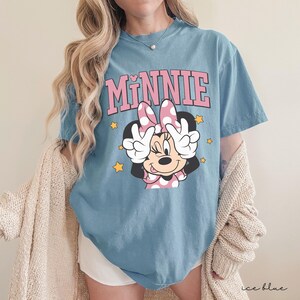 以下が含まれることがあります： ミニーマウスがピースサインをしているグラフィックが特徴のアイスブルーのTシャツ。「MINNIE」の文字がピンク色のブロック体でキャラクターの上にプリントされています。黄色の星がグラフィックの周りに散りばめられています。ゆったりとしたフィット感のシャツです。