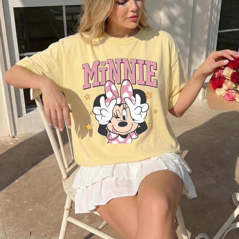 以下が含まれることがあります： ミニーマウスのグラフィックとピンク色の「MINNIE」の文字が入った薄い黄色のTシャツ。ミニーマウスはピンクのドット柄のリボンとドレスを着ています。ゆったりとしたフィット感のTシャツで、白いスカートと合わせています。