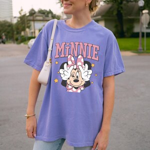 以下が含まれることがあります： ミニーマウスのグラフィックとピンク色の「MINNIE」の文字が入ったライラック色のTシャツ。ミニーは、目の前にピースサインを作り、黄色の星に囲まれています。カジュアルなスタイル。