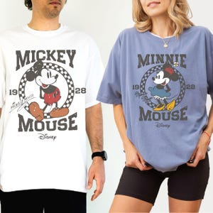 Puede incluir: Dos camisetas con Mickey y Minnie Mouse de Disney. La camiseta blanca tiene a Mickey Mouse en un c&iacute;rculo con el texto "Mickey Mouse 1928". La camiseta azul tiene a Minnie Mouse con el texto "Minnie Mouse 1928".