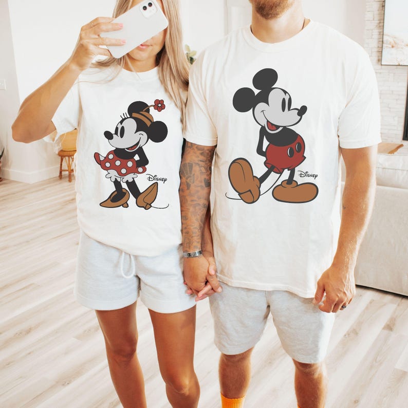 Puede incluir: Camisetas blancas a juego con Mickey y Minnie Mouse. Minnie lleva un vestido de lunares rojos y una flor. Mickey lleva pantalones cortos rojos. Ambas camisetas tienen el logo de Disney.